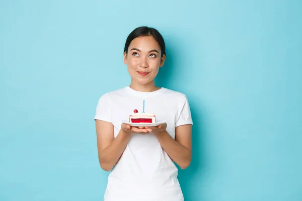 Férias, estilo de vida e conceito de celebração. Retrato de sonho sorrindo menina asiática em branco t-shirt olhando para cima pensativo enquanto segurando pedaço de bolo de aniversário, fazendo desejo na vela acesa — Fotografia de Stock