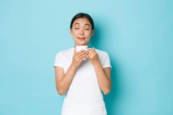 Portrait de ravie rêveuse, jolie fille asiatique en t-shirt blanc, les yeux fermés et le café odorant, profiter de la routine matinale, siroter cappuccino de tasse, debout fond bleu clair — Photo