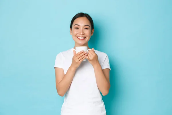 Retrato de sorrir menina asiática feliz em t-shirt branca, olhando satisfeito depois de beber sua caneca da manhã de café, de pé sobre fundo azul claro com sorriso satisfeito — Fotografia de Stock