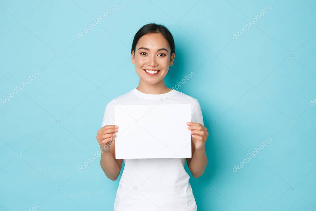 Waist-up portrait of smiling beautiful asian girl searching for someone, making announcement, holding blank piece of paper and looking at camera, standing light blue background