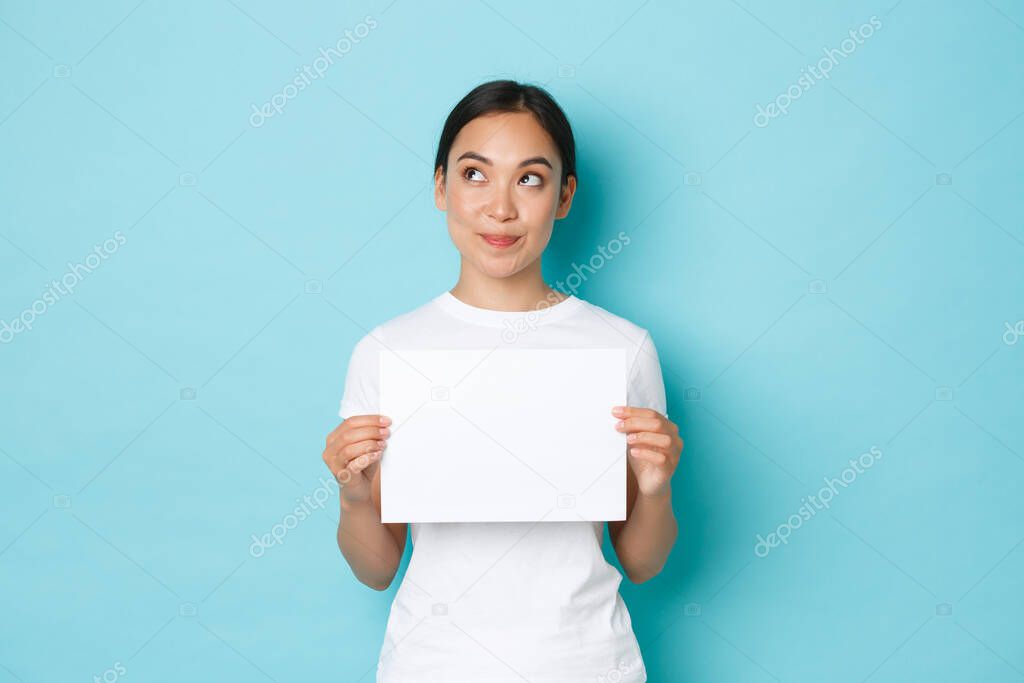 Portrait of thoughtful dreamy, pretty asian female in white t-shirt, looking upper left corner and thinking, holding piece of blank paper, making announcement, pondering over light blue background