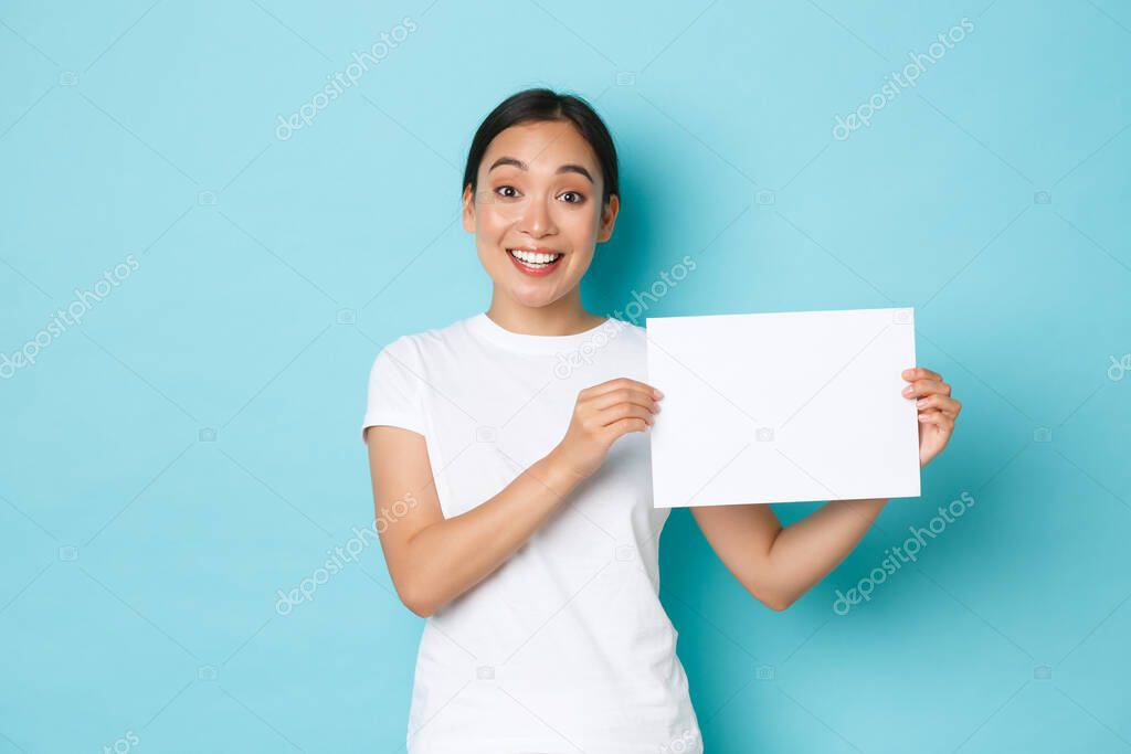 Waist-up portrait of smiling beautiful asian girl searching for someone, making announcement, holding blank piece of paper and looking at camera, standing light blue background