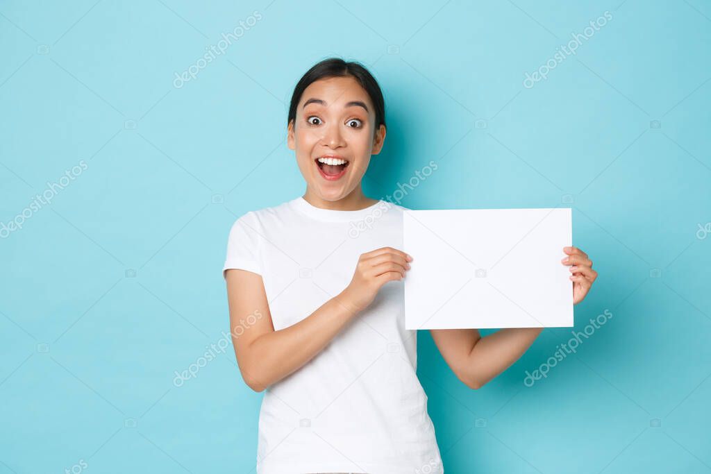 Amazed and excited asian girl in white t-shirt, making announcement, gasping surprised while showing sign, piece of blank paper with your advertisement, standing light blue background