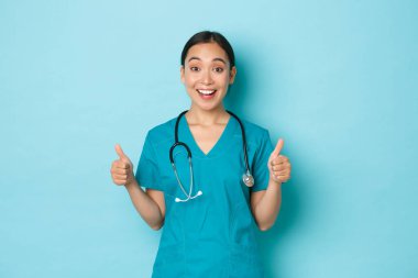 Covid-19, sosyal uzaklık ve koronavirüs salgını konsepti. Heyecanlı ve şaşkın gülümseyen önlüklü Asyalı kadın doktor iyi bir fikir duyup, önlükler ve onaylar içinde ayakta durup, kliniği tavsiye ediyor.