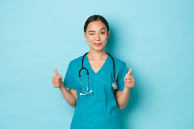 Covid-19, sosyal uzaklık ve koronavirüs salgını konsepti. Şımarık, önlüklü, güzel Asyalı kız kliniğini ziyaret etmeyi teşvik ediyor. Onayladığında onaylıyor ve onaylıyor. Açık mavi arka plan.