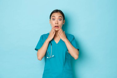 Covid-19, sosyal uzaklık ve koronavirüs salgını konsepti. Şaşırmış, şaşırmış Asyalı doktor, nefes nefese kalmış ve elleri ağzında, açık mavi arka planda heyecanlı görünüyor.