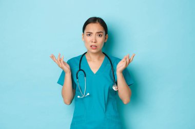 Covid-19, sosyal uzaklık ve koronavirüs salgını konsepti. Kafası karışmış ve rahatsız Asyalı kadın doktor, önlüklü hemşire bir şeyi anlayamıyor, omuz silkip ellerini yana açıyor.