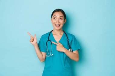 Covid-19, sosyal uzaklık ve koronavirüs salgını konsepti. Neşeli, rüya gibi Asyalı hemşire, önlüklü doktor bilgi ya da tanıtım görüyor, sol üst köşeyi işaret ediyor, açık mavi arka plan