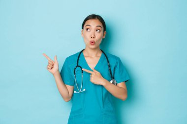 Covid-19, sosyal uzaklık ve koronavirüs salgını konsepti. Şaşırmış ve etkilenmiş Asyalı kadın doktor, doktor ya da hemşire merak ediyor, sol üst köşeyi işaret ediyor.