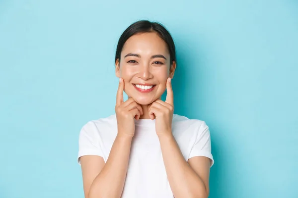 Nahaufnahme der schönen asiatischen Mädchen, die zufrieden aussehen, lächelnd weiße Zähne, berühren Wangen, empfehlen Hautpflegeprodukt, Kosmetik für Gesichtshaut, stehend hellblauer Hintergrund — Stockfoto