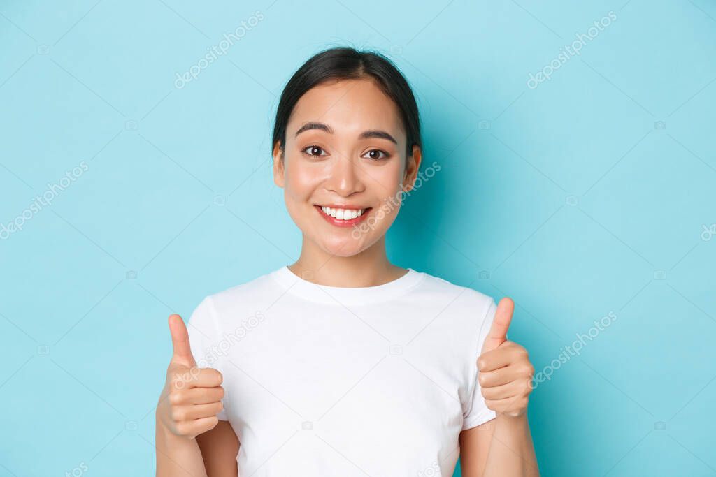Close-up of satisfied and cheerful smiling asian girl in white t-shirt, showing thumbs-up supportive and looking pleased at camera, recommend product, compliment good choice
