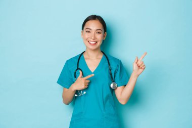 Covid-19, sosyal uzaklık ve koronavirüs salgını konsepti. Neşeli, rüya gibi Asyalı hemşire, önlüklü doktor bilgi ya da tanıtım görüyor, sağ üst köşeyi işaret ediyor, açık mavi arka plan.