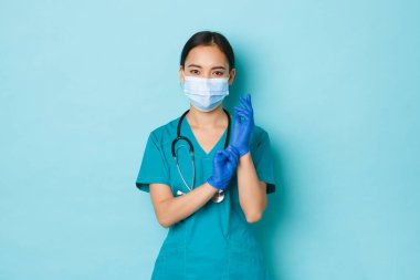 Covid-19, sosyal uzaklık ve koronavirüs salgını konsepti. Kendine güvenen Asyalı doktor, sağlık kontrolü için hazırlanan hemşire, tıbbi maske takan, ciddi bir yüzle lastik eldiven takan.