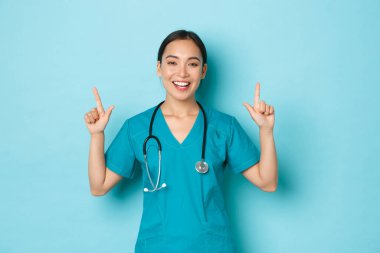 Covid-19, sosyal uzaklık ve koronavirüs salgını konsepti. Gülümseyen çekici Asyalı hemşire, doktor ya da stajyer doktor, parmakla göstererek, büyük haberler vererek, açık mavi arka plan