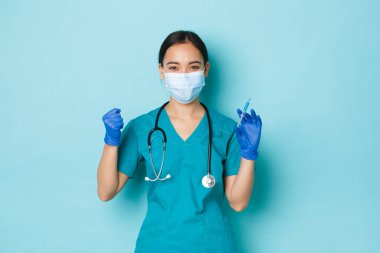 Covid-19, sosyal uzaklık ve koronavirüs salgını konsepti. Kararlı ve neşeli Asyalı kadın doktor, tıbbi maskeli hemşire, eldiven ve aşı şırıngası tutan önlük.