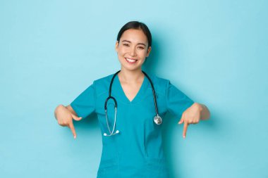 Covid-19, sosyal uzaklık ve koronavirüs salgını konsepti. Hoş, sevimli Asyalı bayan doktor, hemşire reklamını yapıyor, parmaklarını eğiyor ve gülümsüyor, açık mavi arka plan.