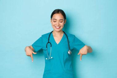 Covid-19, sosyal uzaklık ve koronavirüs salgını konsepti. Çekici Asyalı doktor, laboratuardaki ya da klinikteki hemşire tatmin olmuş bir şekilde aşağıya bakıyor, açık mavi arka planda duruyor.