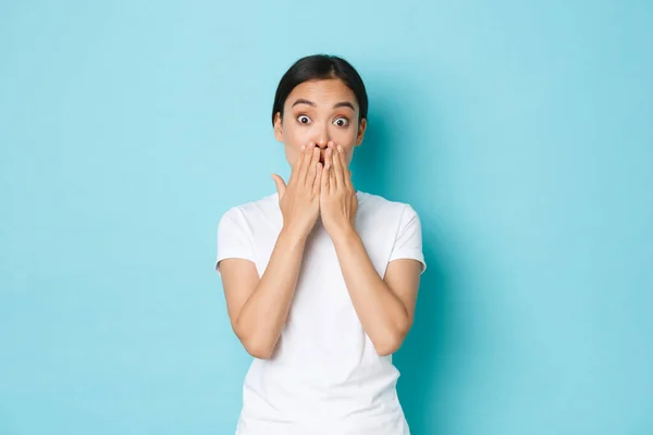 Lifestyle, beauty and shopping concept. Portrait of shocked and amazed, gasping asian female student in casual t-shirt, cover mouth and looking ambushed from shocking news, blue background