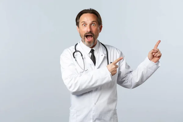 Covid-19, coronavirus outbreak, healthcare workers and pandemic concept. Impressed and excited male doctor in white coat, reacting to wonderful news, pointing upper right corner thrilled — Stock Photo, Image