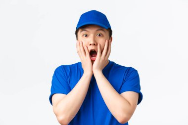 Çevrimiçi alışveriş, hızlı nakliye, çalışanlar ve ev teslimatı konsepti. Şaşırmış ve şaşırmış mavi üniformalı Asyalı erkek kurye, hayret içinde, el ele tutuşmuş, beyaz arka plan