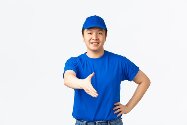 Vriendelijk vrolijke Aziatische koerier in blauw uniform glimlachend zelfverzekerd, hand uit te breiden voor handdruk, begroetende klant, het maken van deal. Bezorger zegt hallo en staande witte achtergrond — Stockfoto