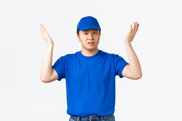 Μπερδεμένος και μπερδεμένος Ασιάτης κούριερ με μπλε στολή δεν καταλαβαίνει, ρωτώντας γιατί, σηκώνοντας τα χέρια ψηλά και κουνώντας τα σαστισμένα, στέκεται ανίδεος πάνω από λευκό φόντο με απογοητευμένη έκφραση — Φωτογραφία Αρχείου