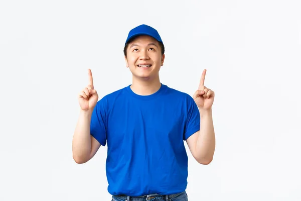 Χαρούμενος Ασιάτης ντελιβεράς με σιδεράκια, φοράει μπλε στολή, δείχνει και κοιτάζει την κορυφαία διαφήμιση, επιδεικνύει το promo προς τα πάνω, χαμογελώντας κατάπληκτος, διαβάζοντας πινακίδα, λευκό φόντο — Φωτογραφία Αρχείου