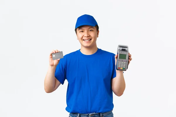 Compras sem contato, pagamento e conceito de funcionários. Sorrindo, mensageiro de aparência amigável, homem de entrega em uniforme azul mostrando cartão de crédito e terminal de POS, de pé fundo branco — Fotografia de Stock