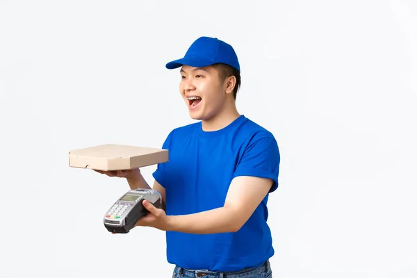 Conceito de compras, pagamento e entrega sem contato. Vista lateral do sorriso agradável asiático mensageiro masculino em uniforme azul trazer pizza para o cliente em casa, dar terminal POS para pagamento rápido, fundo branco — Fotografia de Stock