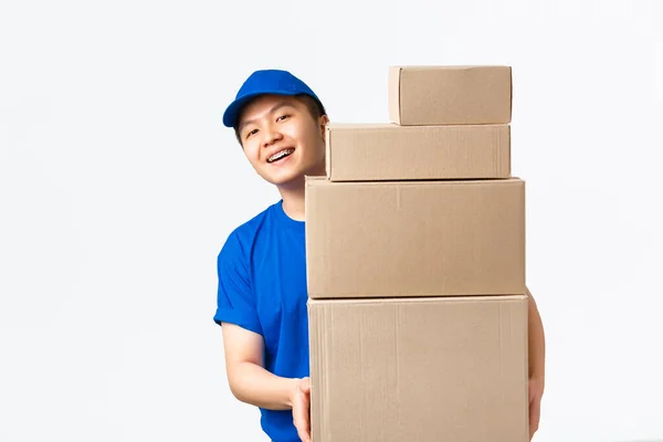 Compras online, concepto de envío rápido. sonriente feliz asiático repartidor hombre en azul mensajero uniforme, mirando detrás de cajas con clientes pedidos, llevar paquetes a casa del cliente, fondo blanco —  Fotos de Stock