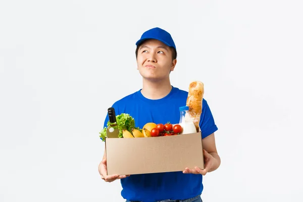 Online shopping, food delivery and shipment concept. Thoughtful young asian male courier in blue uniform, holding box with client order, bring groceries and thinking, looking upper left corner