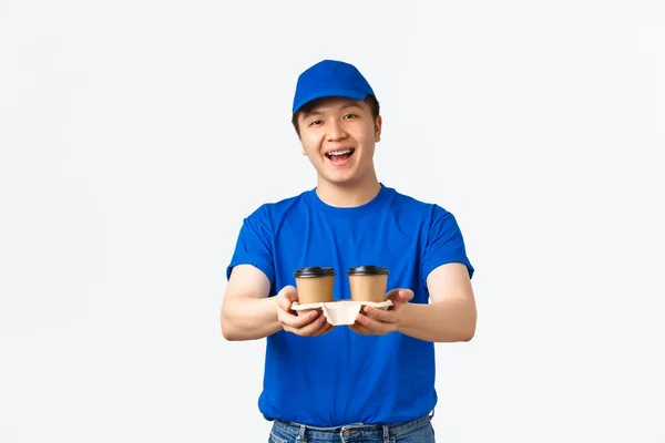 Pedidos en línea, comida para llevar y concepto de empleados. Amistoso hombre de reparto asiático sonriente en uniforme de mensajero azul, la entrega de tazas de café al cliente con expresión alegre, de pie fondo blanco — Foto de Stock