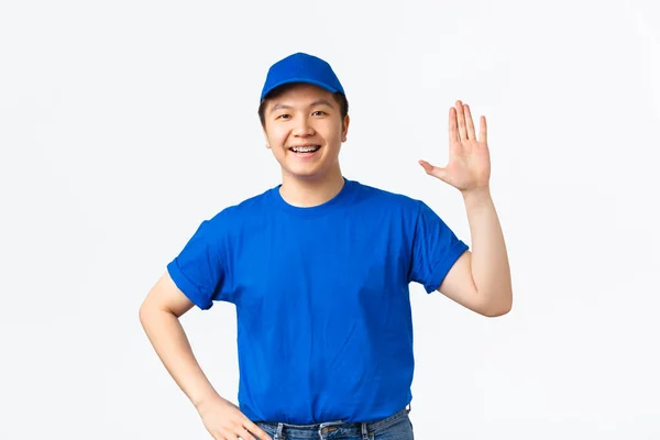 Vriendelijke Aziatische mannelijke koerier in blauw uniform glimlachend en aarzelende hand om hallo te zeggen, groet klant brengen aankoop aan klant deur. Vrolijke bezorger, hi gebaar, witte achtergrond — Stockfoto