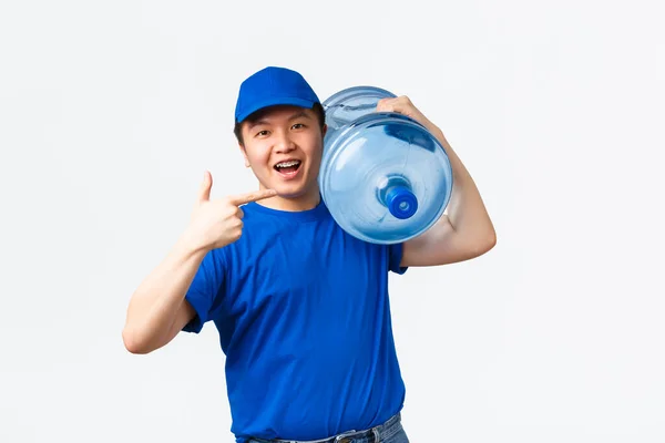 Office and home delivery and employees concept. Young cheerful asain male courier in blue uniform, carry bottle with clean water on shoulder, bring order to client and showing thumbs-up — Stock Photo, Image