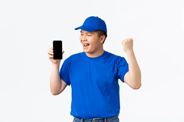 Tecnologia, compras on-line e conceito de transporte. Triunfando alegre asiático entregador homem em azul courier uniforme, mostrando punho bomba e olhando para a tela do telefone móvel, celebrando — Fotografia de Stock