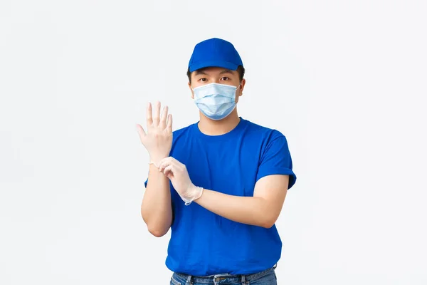Απρόσκοπτη παράδοση, μεταφορά covid-19, πρόληψη της έννοιας του ιού. Νεαρός Ασιάτης υπάλληλος με μπλε στολή κούριερ, φοράει προστατευτικά γάντια πριν παραδώσει πακέτα σε πελάτες, λευκό φόντο — Φωτογραφία Αρχείου