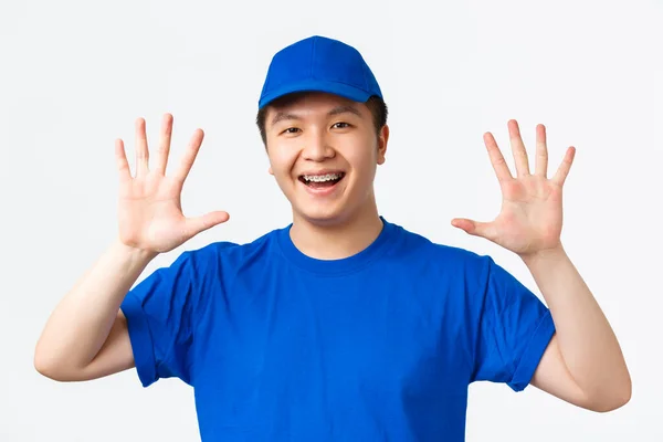 Online-Einkauf, schneller Versand, Mitarbeiter und Lieferkonzept nach Hause. Selbstbewusst lächelnder asiatischer Kurier mit Hosenträgern, in blauer Uniform, Nummer zehn, leere Hände, weißer Hintergrund — Stockfoto
