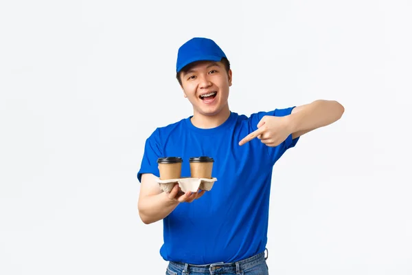 Online orders, takeaway and employees concept. Friendly cheerful male courier in blue uniform ensure fast delivery from restaurants or cafes, pointing finger at coffee and smiling, white background — Stock Photo, Image