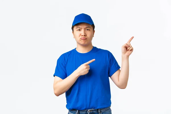 Upprörd och dyster asiatisk kurir i blå mössa och t-shirt grimma disapointed och pekar övre högra hörnet. Sorglig leverans kille i uniform informera dåliga nyheter, stående vit bakgrund — Stockfoto