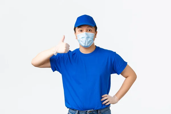 Kontaktlös leverans, covid-19 frakt, förhindra virus koncept. Asiatiska manliga kurir i blå uniform mössa och t-shirt, bär medicinsk mask med handskar, visar tummen upp, garantera säkerheten för paket — Stockfoto