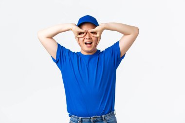 Mavi üniformalı kaygısız gülümseyen Asyalı kurye, süper kahraman maskesi takıyor, el gözlüğü gösteriyor. Beyaz arka planda duran komik teslimatçı. Paketlerin güvende olduğundan emin ol.