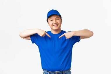 Arkadaşça gülümseyen ve kendine güvenen mavi üniformalı Asyalı teslimatçı kendini işaret ediyor. Kurye şirket hizmetini destekliyor, hızlı ve güvenli nakliye garantisi veriyor, beyaz arka planda duruyor.