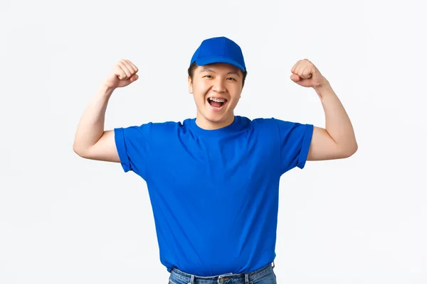 Alegre sonriente asiático entrega hombre flex bíceps y garantizar la seguridad de los paquetes. Mensajero en uniforme azul mostrar sus manos fuertes, llevar paquetes a los hogares de los clientes, de pie fondo blanco — Foto de Stock