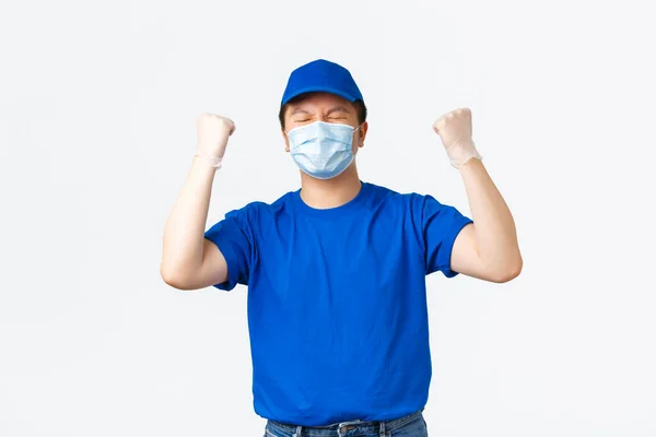 Απρόσκοπτη παράδοση, μεταφορά covid-19, πρόληψη της έννοιας του ιού. Επιτυχημένος ασιατικός κούριερ με μπλε στολή, ιατρική μάσκα και γάντια που χαίρεται, αντλία γροθιάς και λέει ναι — Φωτογραφία Αρχείου
