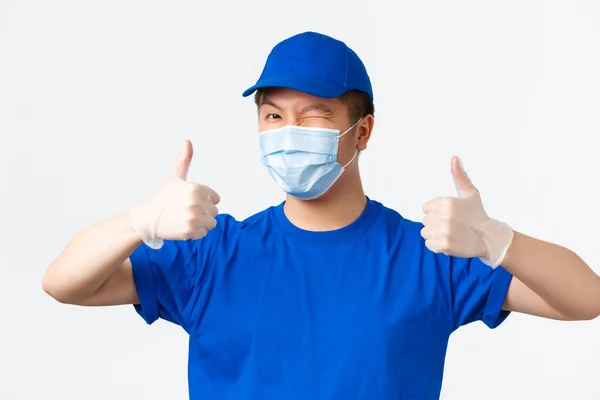 Online αγορές, παράδοση και coronavirus πανδημία, την πρόληψη της έννοιας του ιού. Κοντινό πλάνο του χαρούμενου Ασιάτη κούριερ με μπλε στολή, ιατρική μάσκα και γάντια που δείχνει αντίχειρες σε έγκριση — Φωτογραφία Αρχείου