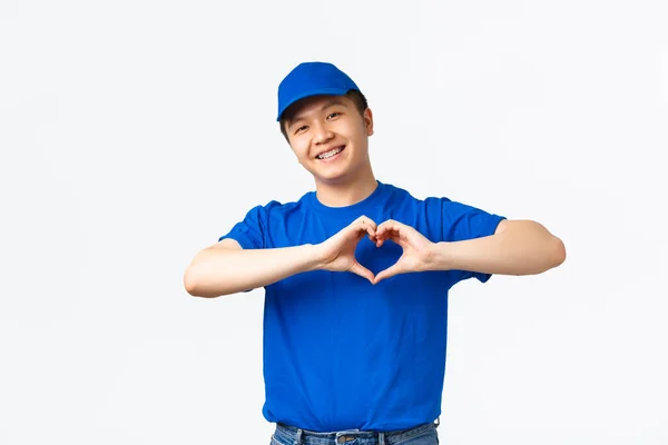 Retrato de carinho amigável asiático entregador homem em azul uniforme valorizar e cuidar de clientes pacotes, mostrando gesto do coração sobre o peito e sorrindo. Courier gosta de seus clientes — Fotografia de Stock