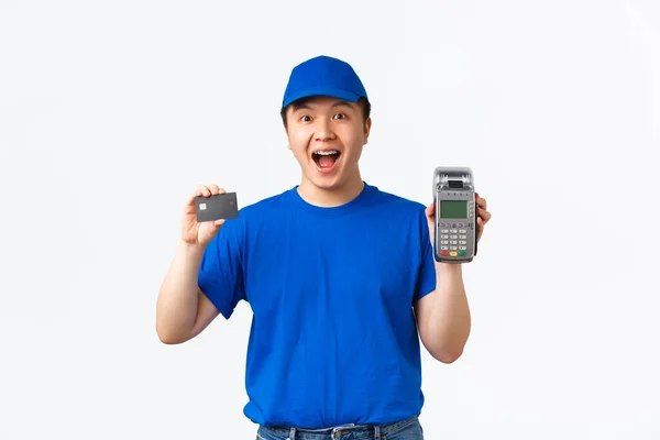 Compras sem contato, pagamento e conceito de funcionários. Alegre feliz asiático mensageiro masculino, homem de entrega em uniforme azul, mostrando cartão de crédito e terminal de POS, pagando fácil e rápido, fundo branco — Fotografia de Stock