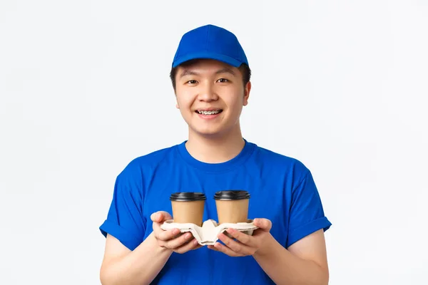 Onlineorder, hämtmat och medarbetarkoncept. Snygg leende asiatisk leverans man i blå kurir uniform, räckte kaffe till kund och ser vänliga, stående vit bakgrund — Stockfoto