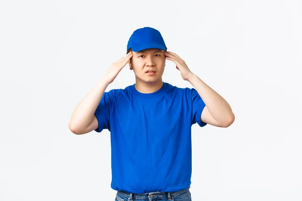 Zűrös ázsiai futár kék sapkában és pólóban szédültnek vagy túlhajszoltnak tűnik. Szállítási srác nem tudja kezelni a nyomást, megható fej, érzés fejfájás vagy fáradt, álló fehér háttér — Stock Fotó
