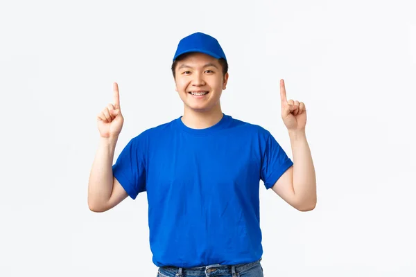 Upbeat leende asiatisk kurir med hängslen, bär blå företagets uniform, peka finger upp, informera kunder om rabatter på sjöfarten, specialerbjudanden, stående vit bakgrund — Stockfoto